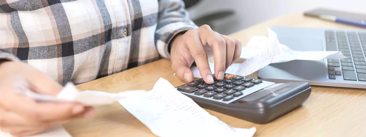 ผู้หญิงกำลังกดเครื่องคิดเลขคำนวณค่าใช้จ่ายต่าง ๆ บนโต๊ะ